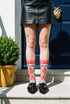 Pair of crew length socks with an all-over folk art print on a cream background. Red anatomical hearts stack vertically between symmetrical floral sprays with fine cream vessel lines, curling green stems and leaves, red flowers, purple tulip-shaped blooms, and small yellow accents. Red cuffs carry cream scrolling vines and small hearts above a scalloped border. Reinforced black heel and toe, striped ribbed cuff. Maximalist medical gift socks.