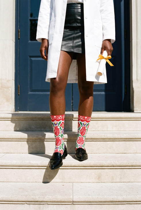 Pair of crew length socks with an all-over folk art print on a cream background. Red anatomical hearts stack vertically between symmetrical floral sprays with fine cream vessel lines, curling green stems and leaves, red flowers, purple tulip-shaped blooms, and small yellow accents. Red cuffs carry cream scrolling vines and small hearts above a scalloped border. Reinforced black heel and toe, striped ribbed cuff. Maximalist medical gift socks.