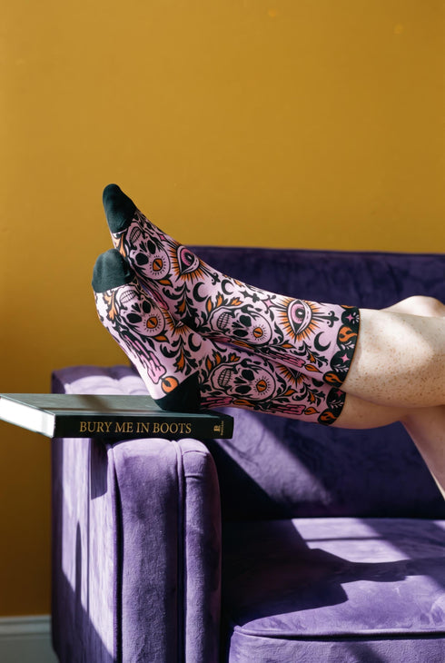 Pair of crew length socks with an all-over pastel goth print on a pastel pink background. Black-outlined human skulls with small orange eyes on their foreheads, all-seeing eyes with orange rays, crescent moons, curling leaves, and pink diamond stars sit in a symmetrical dark damask layout. Black cuff with a scalloped edge, orange flames, and pink flourishes. Reinforced black heel and toe, striped ribbed cuff. Western goth tattoo flash style.