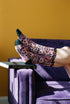 Pair of crew length socks with an all-over pastel goth print on a pastel pink background. Black-outlined human skulls with small orange eyes on their foreheads, all-seeing eyes with orange rays, crescent moons, curling leaves, and pink diamond stars sit in a symmetrical dark damask layout. Black cuff with a scalloped edge, orange flames, and pink flourishes. Reinforced black heel and toe, striped ribbed cuff. Western goth tattoo flash style.