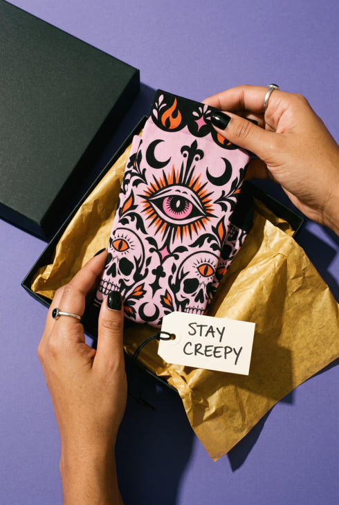 Pair of crew length socks with an all-over pastel goth print on a pastel pink background. Black-outlined human skulls with small orange eyes on their foreheads, all-seeing eyes with orange rays, crescent moons, curling leaves, and pink diamond stars sit in a symmetrical dark damask layout. Black cuff with a scalloped edge, orange flames, and pink flourishes. Reinforced black heel and toe, striped ribbed cuff. Western goth tattoo flash style.