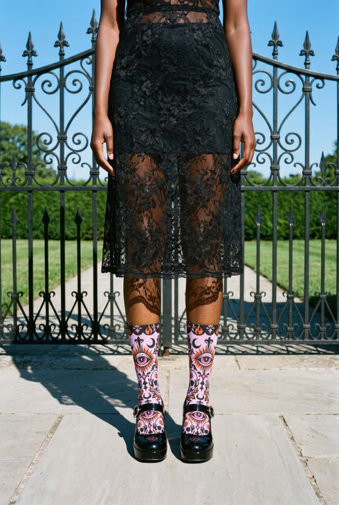 Pair of crew length socks with an all-over pastel goth print on a pastel pink background. Black-outlined human skulls with small orange eyes on their foreheads, all-seeing eyes with orange rays, crescent moons, curling leaves, and pink diamond stars sit in a symmetrical dark damask layout. Black cuff with a scalloped edge, orange flames, and pink flourishes. Reinforced black heel and toe, striped ribbed cuff. Western goth tattoo flash style.