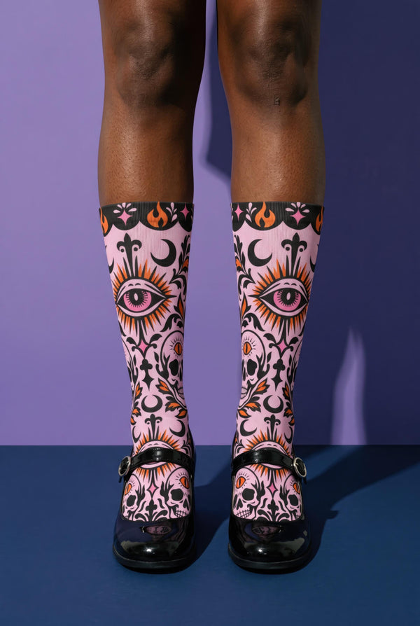 Pair of crew length socks with an all-over pastel goth print on a pastel pink background. Black-outlined human skulls with small orange eyes on their foreheads, all-seeing eyes with orange rays, crescent moons, curling leaves, and pink diamond stars sit in a symmetrical dark damask layout. Black cuff with a scalloped edge, orange flames, and pink flourishes. Reinforced black heel and toe, striped ribbed cuff. Western goth tattoo flash style.