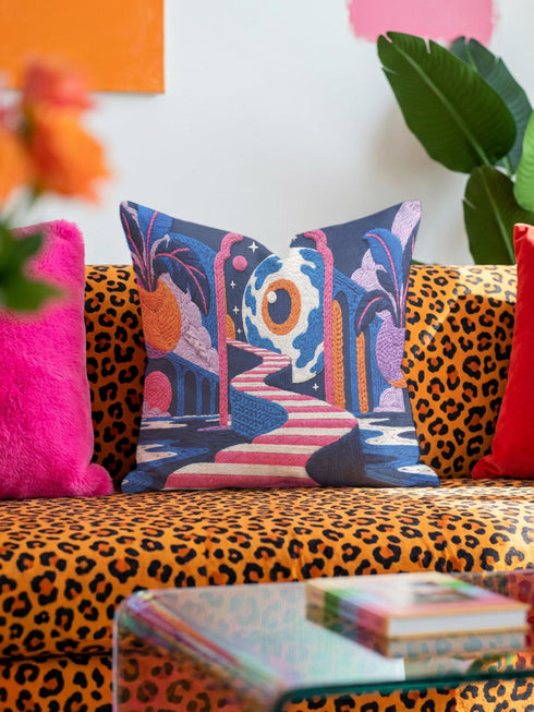 Square surreal liminal space throw pillow cover printed to look like textured embroidery on a dark navy background. A giant blue and white eyeball planet with an orange iris glows inside pink stone arches above a pink and white striped staircase curving over dark blue water, with lavender clouds, blue aqueduct arches, and orange potted palms. Smooth flat 2D print, not raised stitching. Psychedelic dreamscape decor for a sofa, bedroom chair, or reading nook.