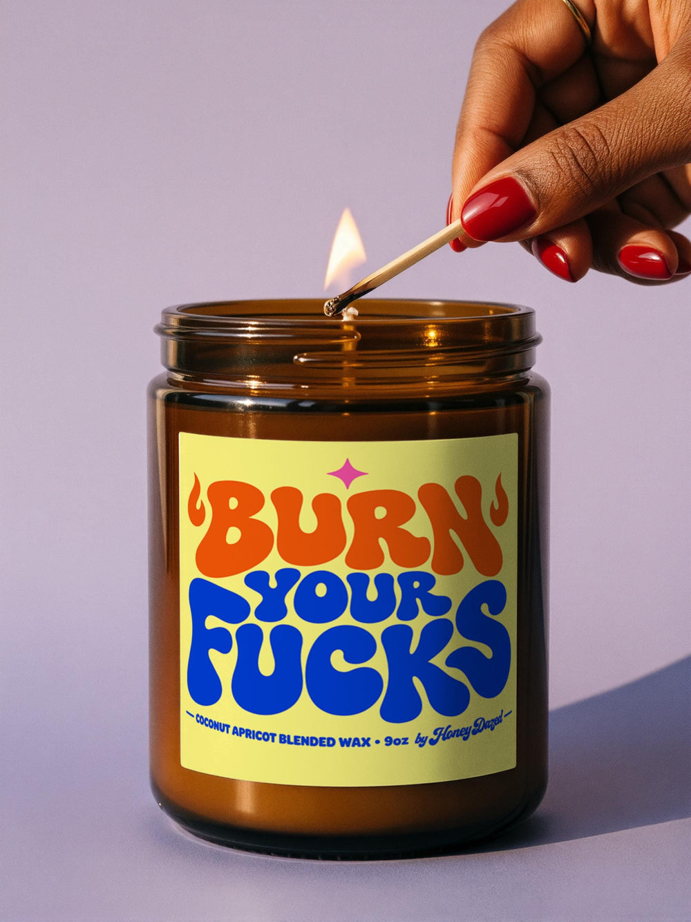 Funny Burn Your Fucks scented candle in a 9 oz amber glass jar with a brushed gold screw lid. Square butter yellow label with 70s retro psychedelic bubble lettering: BURN in orange with groovy flames, YOUR in cobalt blue, FUCKS in big bubbly type under a hot magenta sparkle star, coconut apricot wax 9oz by Honey Dazed along the bottom. Snarky dopamine decor made for an office desk, bathtub ledge, or nightstand, a sweary coworker gift.