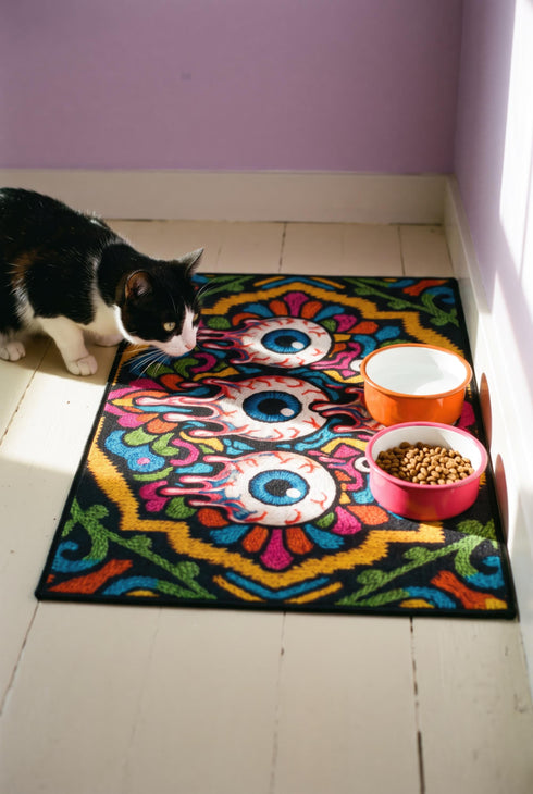 Rectangular doormat with a black background and black edge trim, printed in a flat 2D faux embroidery style. A golden yellow stepped octagon frames four large bloodshot eyeballs with blue irises and dripping pink, with two small peeking eyes among the florals. Hot pink, orange, turquoise, and green folk art petals and green vines fill the corners. Polyester surface with a low pile and non-slip rubber backing. Psychedelic 70s creepy cute decor.