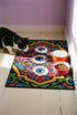 Rectangular doormat with a black background and black edge trim, printed in a flat 2D faux embroidery style. A golden yellow stepped octagon frames four large bloodshot eyeballs with blue irises and dripping pink, with two small peeking eyes among the florals. Hot pink, orange, turquoise, and green folk art petals and green vines fill the corners. Polyester surface with a low pile and non-slip rubber backing. Psychedelic 70s creepy cute decor.