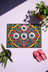 Rectangular doormat with a black background and black edge trim, printed in a flat 2D faux embroidery style. A golden yellow stepped octagon frames four large bloodshot eyeballs with blue irises and dripping pink, with two small peeking eyes among the florals. Hot pink, orange, turquoise, and green folk art petals and green vines fill the corners. Polyester surface with a low pile and non-slip rubber backing. Psychedelic 70s creepy cute decor.
