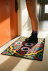 Rectangular doormat with a black background and black edge trim, printed in a flat 2D faux embroidery style. A golden yellow stepped octagon frames four large bloodshot eyeballs with blue irises and dripping pink, with two small peeking eyes among the florals. Hot pink, orange, turquoise, and green folk art petals and green vines fill the corners. Polyester surface with a low pile and non-slip rubber backing. Psychedelic 70s creepy cute decor.