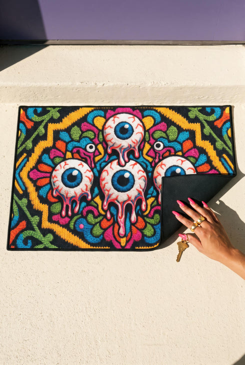 Rectangular doormat with a black background and black edge trim, printed in a flat 2D faux embroidery style. A golden yellow stepped octagon frames four large bloodshot eyeballs with blue irises and dripping pink, with two small peeking eyes among the florals. Hot pink, orange, turquoise, and green folk art petals and green vines fill the corners. Polyester surface with a low pile and non-slip rubber backing. Psychedelic 70s creepy cute decor.