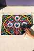 Rectangular doormat with a black background and black edge trim, printed in a flat 2D faux embroidery style. A golden yellow stepped octagon frames four large bloodshot eyeballs with blue irises and dripping pink, with two small peeking eyes among the florals. Hot pink, orange, turquoise, and green folk art petals and green vines fill the corners. Polyester surface with a low pile and non-slip rubber backing. Psychedelic 70s creepy cute decor.