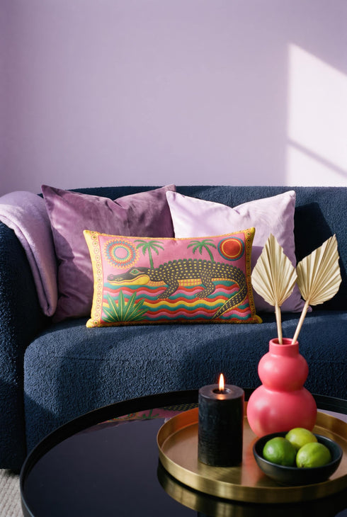 Rectangular 20 by 12 inch lumbar pillow cover with a folk art alligator design printed to look like heavy crewel embroidery on a bright pink background. A large grey and yellow alligator lounges above red, teal, and yellow wavy water lines, flanked by two green palm trees, a pink and yellow sunburst, a red sun, and a green tropical plant. Smooth flat 2D print, not raised stitching. Tropical maximalist decor for a bed, sofa, or reading nook.