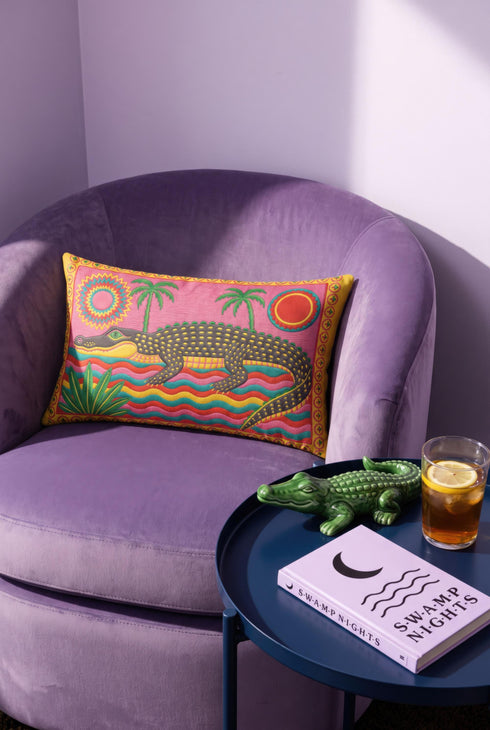 Rectangular 20 by 12 inch lumbar pillow cover with a folk art alligator design printed to look like heavy crewel embroidery on a bright pink background. A large grey and yellow alligator lounges above red, teal, and yellow wavy water lines, flanked by two green palm trees, a pink and yellow sunburst, a red sun, and a green tropical plant. Smooth flat 2D print, not raised stitching. Tropical maximalist decor for a bed, sofa, or reading nook.