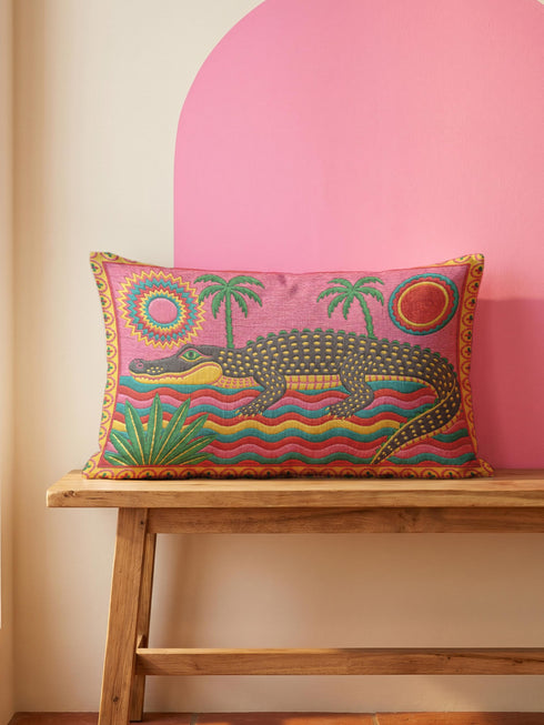 Rectangular 20 by 12 inch lumbar pillow cover with a folk art alligator design printed to look like heavy crewel embroidery on a bright pink background. A large grey and yellow alligator lounges above red, teal, and yellow wavy water lines, flanked by two green palm trees, a pink and yellow sunburst, a red sun, and a green tropical plant. Smooth flat 2D print, not raised stitching. Tropical maximalist decor for a bed, sofa, or reading nook.
