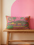 Rectangular 20 by 12 inch lumbar pillow cover with a folk art alligator design printed to look like heavy crewel embroidery on a bright pink background. A large grey and yellow alligator lounges above red, teal, and yellow wavy water lines, flanked by two green palm trees, a pink and yellow sunburst, a red sun, and a green tropical plant. Smooth flat 2D print, not raised stitching. Tropical maximalist decor for a bed, sofa, or reading nook.
