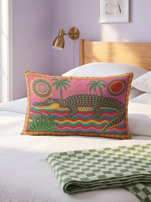 Rectangular 20 by 12 inch lumbar pillow cover with a folk art alligator design printed to look like heavy crewel embroidery on a bright pink background. A large grey and yellow alligator lounges above red, teal, and yellow wavy water lines, flanked by two green palm trees, a pink and yellow sunburst, a red sun, and a green tropical plant. Smooth flat 2D print, not raised stitching. Tropical maximalist decor for a bed, sofa, or reading nook.