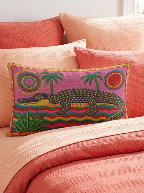 Rectangular 20 by 12 inch lumbar pillow cover with a folk art alligator design printed to look like heavy crewel embroidery on a bright pink background. A large grey and yellow alligator lounges above red, teal, and yellow wavy water lines, flanked by two green palm trees, a pink and yellow sunburst, a red sun, and a green tropical plant. Smooth flat 2D print, not raised stitching. Tropical maximalist decor for a bed, sofa, or reading nook.