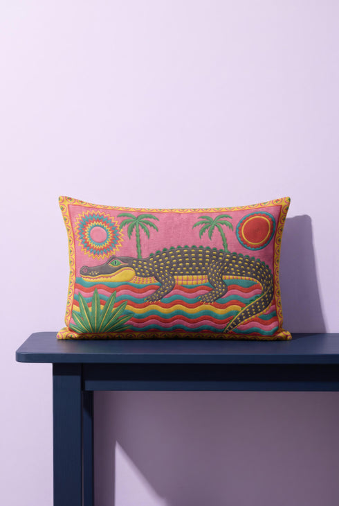 Rectangular 20 by 12 inch lumbar pillow cover with a folk art alligator design printed to look like heavy crewel embroidery on a bright pink background. A large grey and yellow alligator lounges above red, teal, and yellow wavy water lines, flanked by two green palm trees, a pink and yellow sunburst, a red sun, and a green tropical plant. Smooth flat 2D print, not raised stitching. Tropical maximalist decor for a bed, sofa, or reading nook.