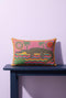 Rectangular 20 by 12 inch lumbar pillow cover with a folk art alligator design printed to look like heavy crewel embroidery on a bright pink background. A large grey and yellow alligator lounges above red, teal, and yellow wavy water lines, flanked by two green palm trees, a pink and yellow sunburst, a red sun, and a green tropical plant. Smooth flat 2D print, not raised stitching. Tropical maximalist decor for a bed, sofa, or reading nook.