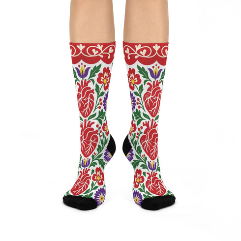 Pair of crew length socks with an all-over folk art print on a cream background. Red anatomical hearts stack vertically between symmetrical floral sprays with fine cream vessel lines, curling green stems and leaves, red flowers, purple tulip-shaped blooms, and small yellow accents. Red cuffs carry cream scrolling vines and small hearts above a scalloped border. Reinforced black heel and toe, striped ribbed cuff. Maximalist medical gift socks.
