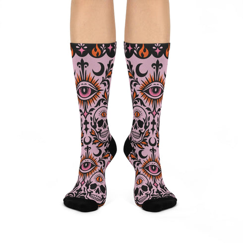 Pair of crew length socks with an all-over pastel goth print on a pastel pink background. Black-outlined human skulls with small orange eyes on their foreheads, all-seeing eyes with orange rays, crescent moons, curling leaves, and pink diamond stars sit in a symmetrical dark damask layout. Black cuff with a scalloped edge, orange flames, and pink flourishes. Reinforced black heel and toe, striped ribbed cuff. Western goth tattoo flash style.