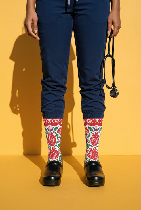 Pair of crew length socks with an all-over folk art print on a cream background. Red anatomical hearts stack vertically between symmetrical floral sprays with fine cream vessel lines, curling green stems and leaves, red flowers, purple tulip-shaped blooms, and small yellow accents. Red cuffs carry cream scrolling vines and small hearts above a scalloped border. Reinforced black heel and toe, striped ribbed cuff. Maximalist medical gift socks.