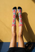 Pair of crew length socks with an all-over folk art print on a cream background. Red anatomical hearts stack vertically between symmetrical floral sprays with fine cream vessel lines, curling green stems and leaves, red flowers, purple tulip-shaped blooms, and small yellow accents. Red cuffs carry cream scrolling vines and small hearts above a scalloped border. Reinforced black heel and toe, striped ribbed cuff. Maximalist medical gift socks.