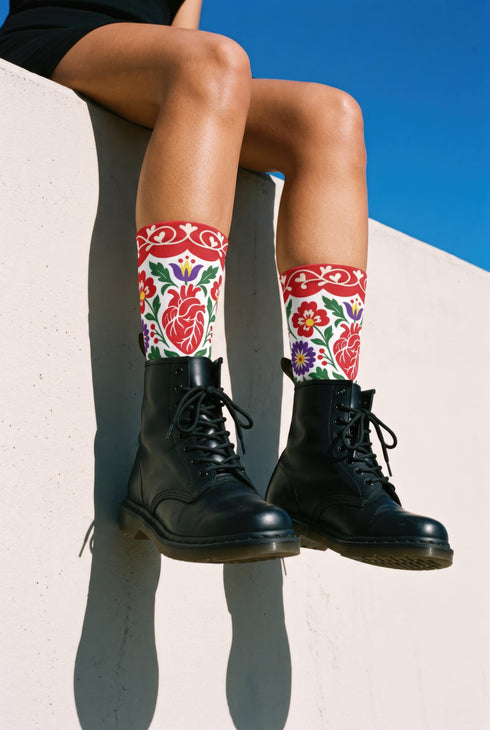 Pair of crew length socks with an all-over folk art print on a cream background. Red anatomical hearts stack vertically between symmetrical floral sprays with fine cream vessel lines, curling green stems and leaves, red flowers, purple tulip-shaped blooms, and small yellow accents. Red cuffs carry cream scrolling vines and small hearts above a scalloped border. Reinforced black heel and toe, striped ribbed cuff. Maximalist medical gift socks.