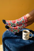 Pair of crew length socks with an all-over folk art print on a cream background. Red anatomical hearts stack vertically between symmetrical floral sprays with fine cream vessel lines, curling green stems and leaves, red flowers, purple tulip-shaped blooms, and small yellow accents. Red cuffs carry cream scrolling vines and small hearts above a scalloped border. Reinforced black heel and toe, striped ribbed cuff. Maximalist medical gift socks.