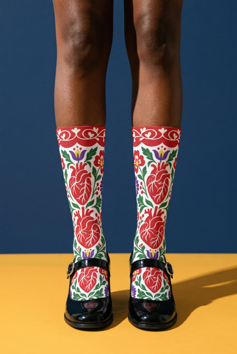 Pair of crew length socks with an all-over folk art print on a cream background. Red anatomical hearts stack vertically between symmetrical floral sprays with fine cream vessel lines, curling green stems and leaves, red flowers, purple tulip-shaped blooms, and small yellow accents. Red cuffs carry cream scrolling vines and small hearts above a scalloped border. Reinforced black heel and toe, striped ribbed cuff. Maximalist medical gift socks.