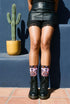 Pair of crew length socks with an all-over pastel goth print on a pastel pink background. Black-outlined human skulls with small orange eyes on their foreheads, all-seeing eyes with orange rays, crescent moons, curling leaves, and pink diamond stars sit in a symmetrical dark damask layout. Black cuff with a scalloped edge, orange flames, and pink flourishes. Reinforced black heel and toe, striped ribbed cuff. Western goth tattoo flash style.