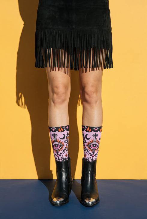 Pair of crew length socks with an all-over pastel goth print on a pastel pink background. Black-outlined human skulls with small orange eyes on their foreheads, all-seeing eyes with orange rays, crescent moons, curling leaves, and pink diamond stars sit in a symmetrical dark damask layout. Black cuff with a scalloped edge, orange flames, and pink flourishes. Reinforced black heel and toe, striped ribbed cuff. Western goth tattoo flash style.