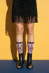 Pair of crew length socks with an all-over pastel goth print on a pastel pink background. Black-outlined human skulls with small orange eyes on their foreheads, all-seeing eyes with orange rays, crescent moons, curling leaves, and pink diamond stars sit in a symmetrical dark damask layout. Black cuff with a scalloped edge, orange flames, and pink flourishes. Reinforced black heel and toe, striped ribbed cuff. Western goth tattoo flash style.