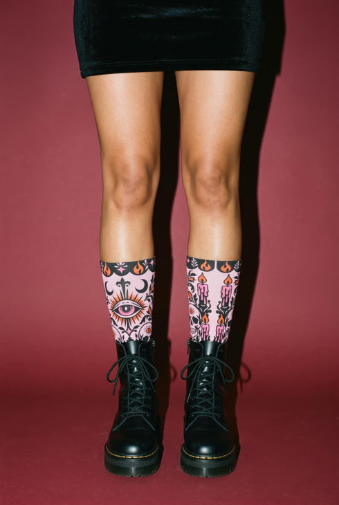 Pair of crew length socks with an all-over pastel goth print on a pastel pink background. Black-outlined human skulls with small orange eyes on their foreheads, all-seeing eyes with orange rays, crescent moons, curling leaves, and pink diamond stars sit in a symmetrical dark damask layout. Black cuff with a scalloped edge, orange flames, and pink flourishes. Reinforced black heel and toe, striped ribbed cuff. Western goth tattoo flash style.