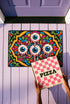 Rectangular doormat with a black background and black edge trim, printed in a flat 2D faux embroidery style. A golden yellow stepped octagon frames four large bloodshot eyeballs with blue irises and dripping pink, with two small peeking eyes among the florals. Hot pink, orange, turquoise, and green folk art petals and green vines fill the corners. Polyester surface with a low pile and non-slip rubber backing. Psychedelic 70s creepy cute decor.