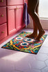 Rectangular doormat with a black background and black edge trim, printed in a flat 2D faux embroidery style. A golden yellow stepped octagon frames four large bloodshot eyeballs with blue irises and dripping pink, with two small peeking eyes among the florals. Hot pink, orange, turquoise, and green folk art petals and green vines fill the corners. Polyester surface with a low pile and non-slip rubber backing. Psychedelic 70s creepy cute decor.