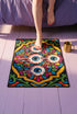Rectangular doormat with a black background and black edge trim, printed in a flat 2D faux embroidery style. A golden yellow stepped octagon frames four large bloodshot eyeballs with blue irises and dripping pink, with two small peeking eyes among the florals. Hot pink, orange, turquoise, and green folk art petals and green vines fill the corners. Polyester surface with a low pile and non-slip rubber backing. Psychedelic 70s creepy cute decor.