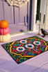 Rectangular doormat with a black background and black edge trim, printed in a flat 2D faux embroidery style. A golden yellow stepped octagon frames four large bloodshot eyeballs with blue irises and dripping pink, with two small peeking eyes among the florals. Hot pink, orange, turquoise, and green folk art petals and green vines fill the corners. Polyester surface with a low pile and non-slip rubber backing. Psychedelic 70s creepy cute decor.