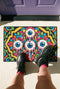 Rectangular doormat with a black background and black edge trim, printed in a flat 2D faux embroidery style. A golden yellow stepped octagon frames four large bloodshot eyeballs with blue irises and dripping pink, with two small peeking eyes among the florals. Hot pink, orange, turquoise, and green folk art petals and green vines fill the corners. Polyester surface with a low pile and non-slip rubber backing. Psychedelic 70s creepy cute decor.