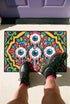 Rectangular doormat with a black background and black edge trim, printed in a flat 2D faux embroidery style. A golden yellow stepped octagon frames four large bloodshot eyeballs with blue irises and dripping pink, with two small peeking eyes among the florals. Hot pink, orange, turquoise, and green folk art petals and green vines fill the corners. Polyester surface with a low pile and non-slip rubber backing. Psychedelic 70s creepy cute decor.