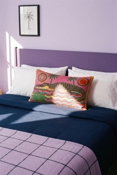 Rectangular 20 by 12 inch lumbar pillow cover with a folk art alligator design printed to look like heavy crewel embroidery on a bright pink background. A large grey and yellow alligator lounges above red, teal, and yellow wavy water lines, flanked by two green palm trees, a pink and yellow sunburst, a red sun, and a green tropical plant. Smooth flat 2D print, not raised stitching. Tropical maximalist decor for a bed, sofa, or reading nook.