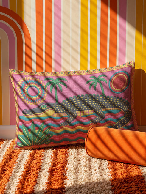Rectangular 20 by 12 inch lumbar pillow cover with a folk art alligator design printed to look like heavy crewel embroidery on a bright pink background. A large grey and yellow alligator lounges above red, teal, and yellow wavy water lines, flanked by two green palm trees, a pink and yellow sunburst, a red sun, and a green tropical plant. Smooth flat 2D print, not raised stitching. Tropical maximalist decor for a bed, sofa, or reading nook.