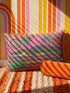 Rectangular 20 by 12 inch lumbar pillow cover with a folk art alligator design printed to look like heavy crewel embroidery on a bright pink background. A large grey and yellow alligator lounges above red, teal, and yellow wavy water lines, flanked by two green palm trees, a pink and yellow sunburst, a red sun, and a green tropical plant. Smooth flat 2D print, not raised stitching. Tropical maximalist decor for a bed, sofa, or reading nook.