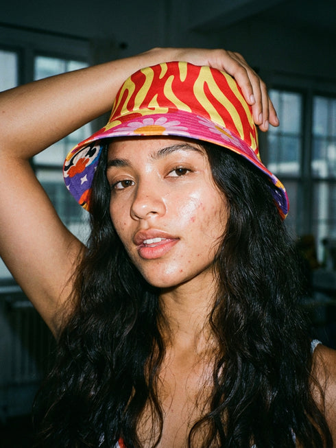 A reversible bucket hat with two distinct sides. Side one features a red and yellow zebra pattern with large pink and orange daisies. Side two has a deep purple background with white stars, rainbow scalloped bands in blue, green, and orange, and retro black and white flowers. The fabric has a visible textured linen weave. The hat is shown in a structured, professional studio shot against a white background, highlighting its 100% polyester durability.
