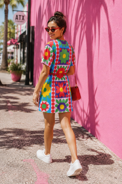 Short-sleeve crew-neck t-shirt dress falling to mid-thigh, with a bold retro patchwork floral all-over print. Large mod daisies in red, purple, yellow, and green sit across a sky blue background, with a central column framing single flowers on purple and pink tiles. Red and pink beaded borders run down the front, and green leaves and more flowers cover the sleeves. The relaxed pull-on dress has a flat printed surface in vivid, high-contrast color.