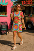 Short sleeve crew neck t-shirt dress on a cream background with an allover retro mod flower medallion print. Bold 60s daisies in red, yellow, and white sit at the center of concentric rainbow rings in lime green, hot pink, orange, and royal blue, with small magenta squares and tiny red checkerboard accents between the motifs. Above-the-knee straight fit silhouette in a 95 percent cotton, 5 percent elastane heavyweight stretch blend. Groovy retro flower power dress for dopamine dressing and easy summer outfi