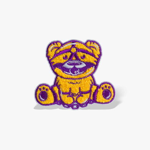 Patch featuring a cute bear in yellow and purple with a playful expression, wearing a BDSM harness and a ball gag. The design is fun and whimsical, combining bold colors with a cheeky style.