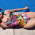 A colorful one-piece swimsuit features a vibrant, quirky pattern with cherries, cartoon eyes, and playful designs on a dark background.
