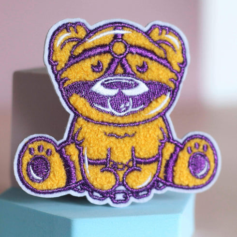 Patch featuring a cute bear in yellow and purple with a playful expression, wearing a BDSM harness and a ball gag. The design is fun and whimsical, combining bold colors with a cheeky style.