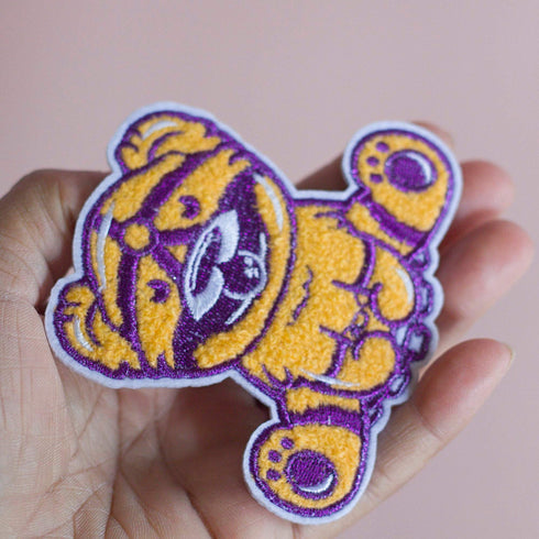Patch featuring a cute bear in yellow and purple with a playful expression, wearing a BDSM harness and a ball gag. The design is fun and whimsical, combining bold colors with a cheeky style.